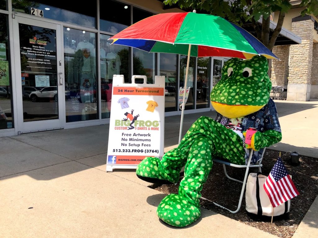 A Big Frog Franchise Mascot, Sitting In A Chair, Under An Umbrella.